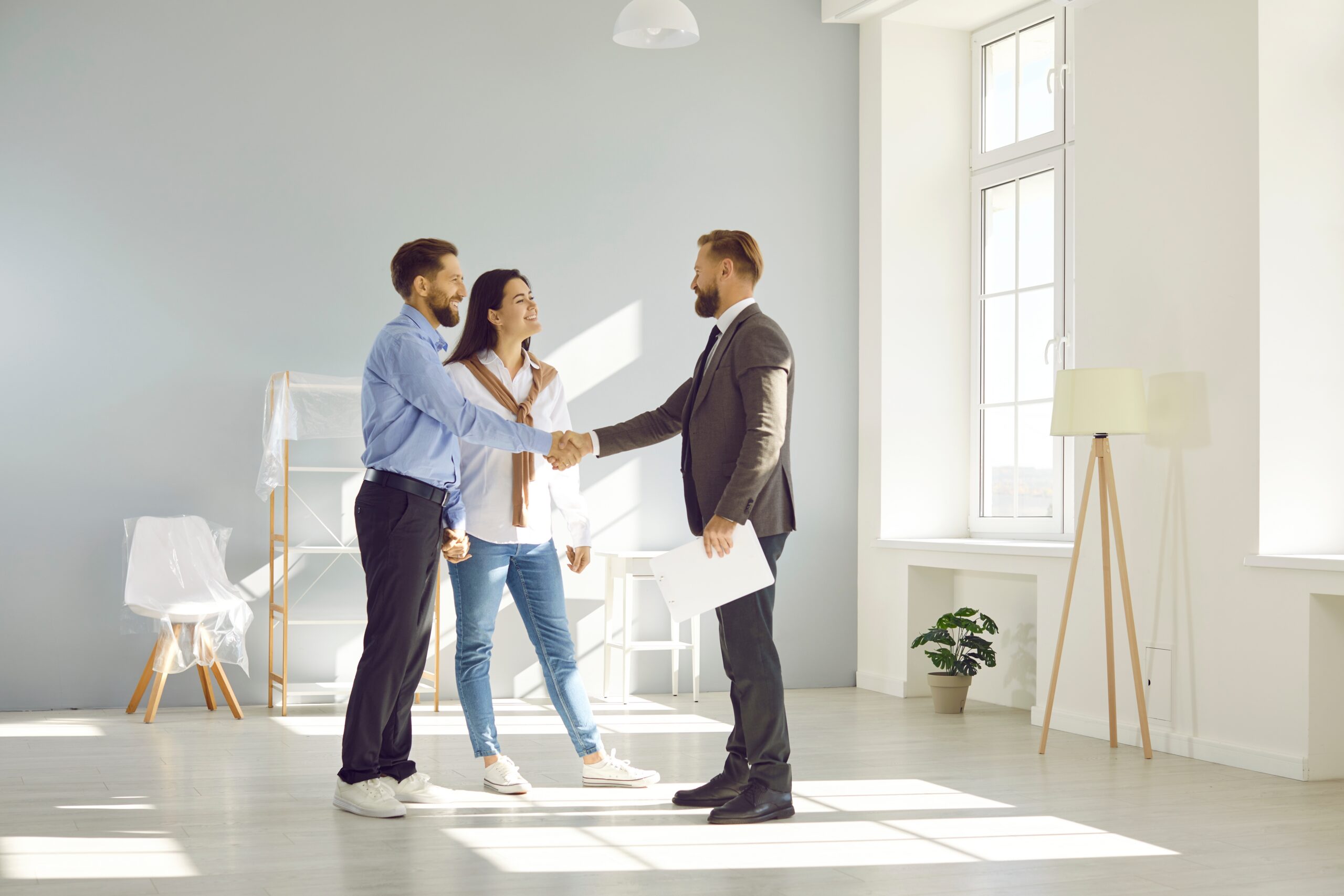 Young married couple smiling and shaking hands after finalizing the purchase of their new home with a real estate agent.
